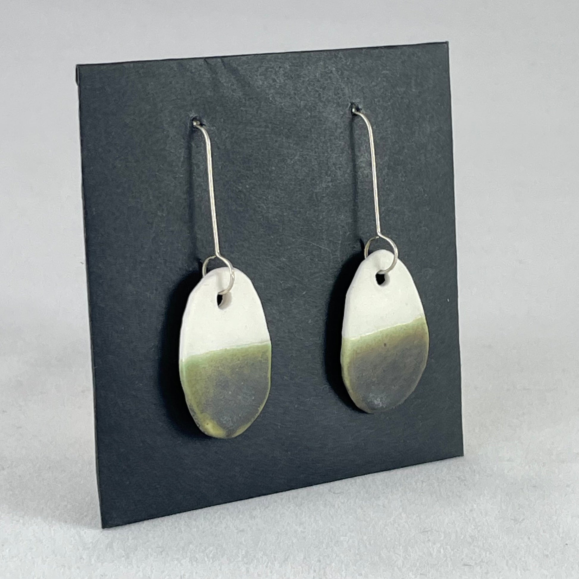 Side view of a pair of oval shaped porcelain earrings in white and green. On sterling silver earrings hooks.