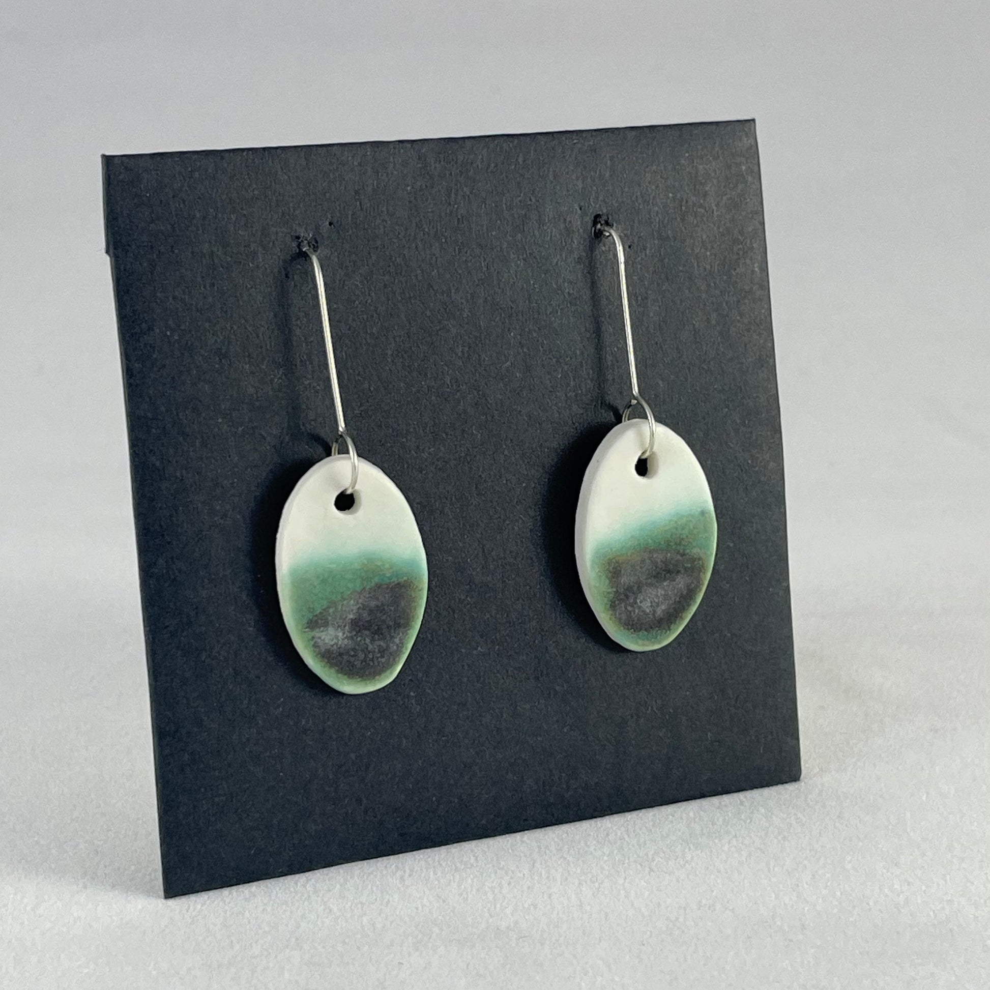 Pair of emerald green and white oval shaped porcelain earrings. Top half white and bottom half textured green. Sterling silver hooks. Side view