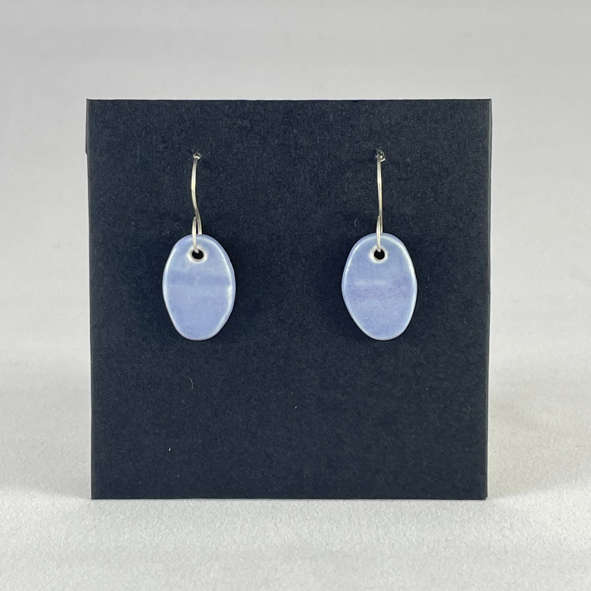 A pair of oval shaped porcelain earrings in blue. Photographed on a black background. On sterling silver earrings hooks.