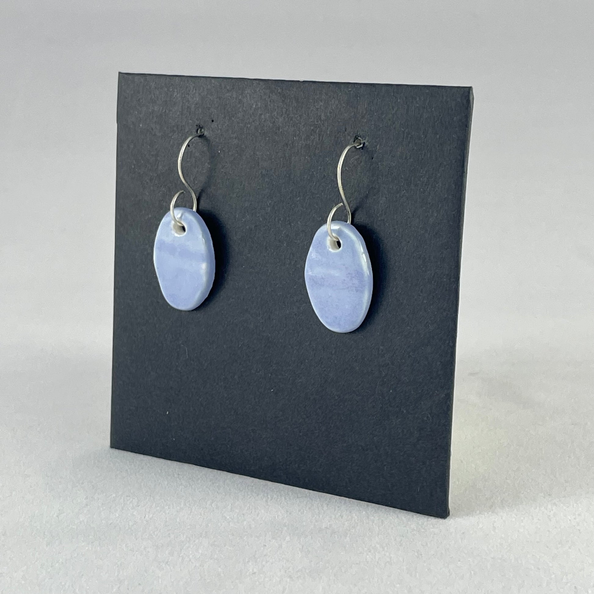 A pair of oval shaped porcelain earrings in blue. Photographed on a black background. On sterling silver earrings hooks. Side view
