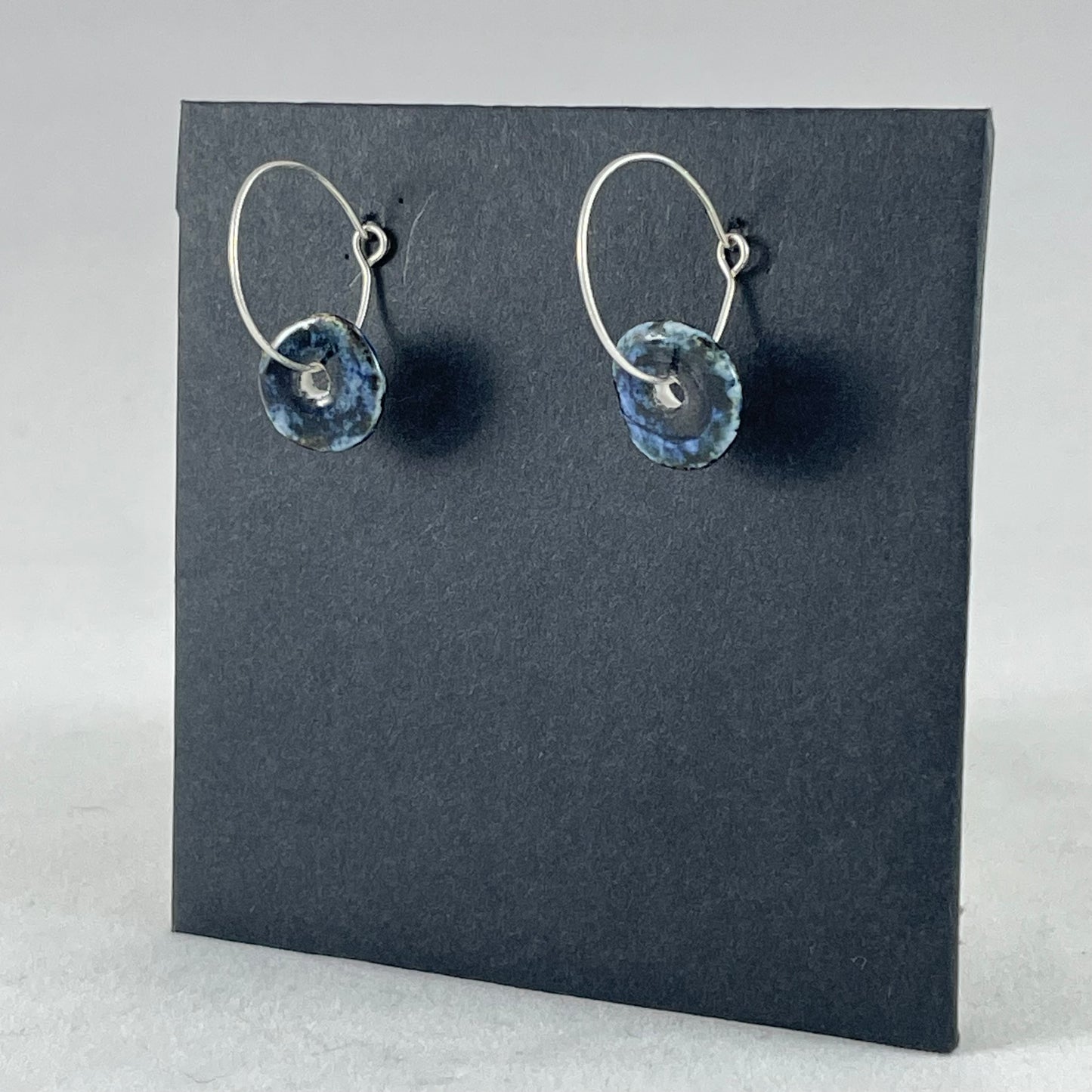 A pair of blue hand coiled porcelain earrings on sterling silver hoops. Photographed on a graphite coloured background. Side view