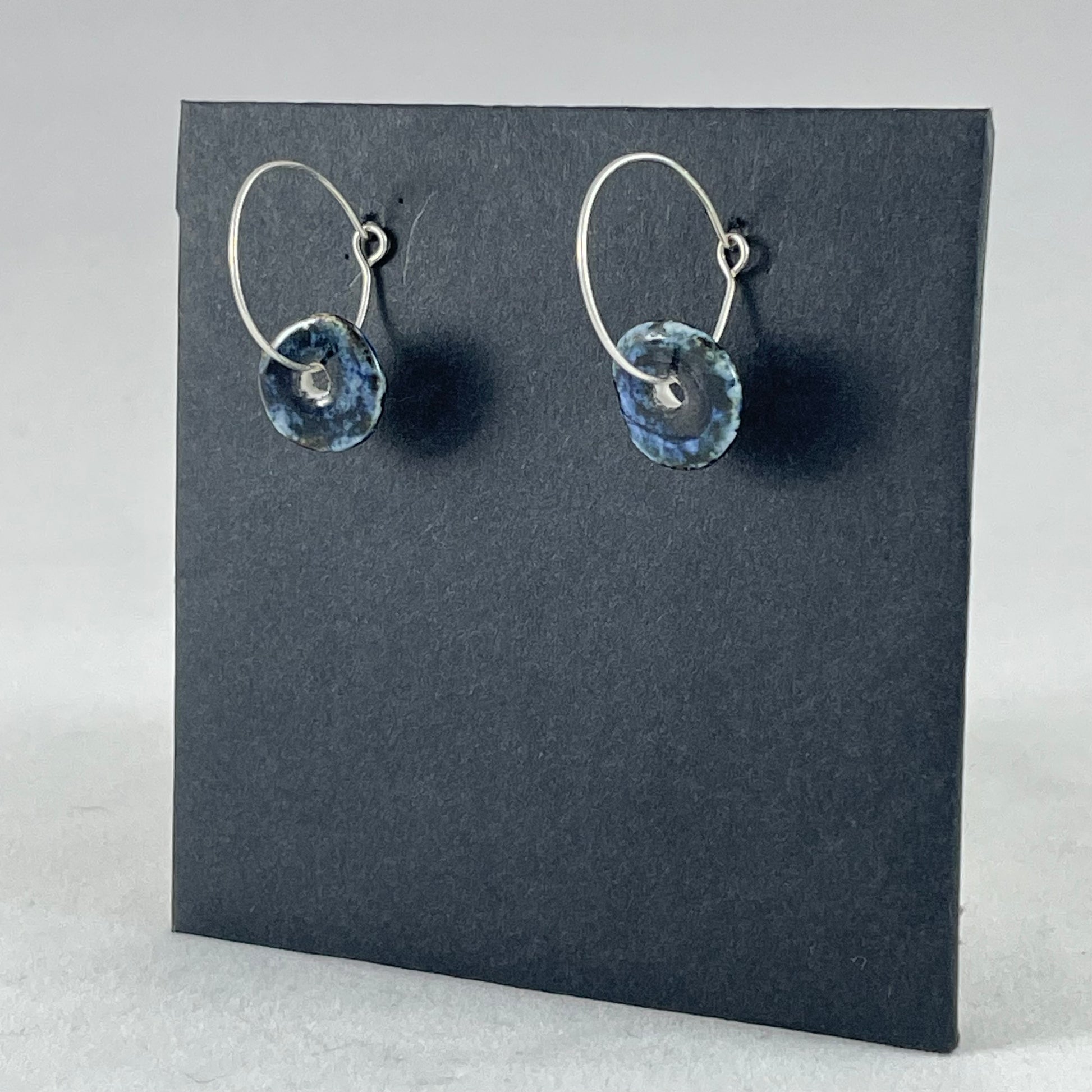 A pair of blue hand coiled porcelain earrings on sterling silver hoops. Photographed on a graphite coloured background. Side view