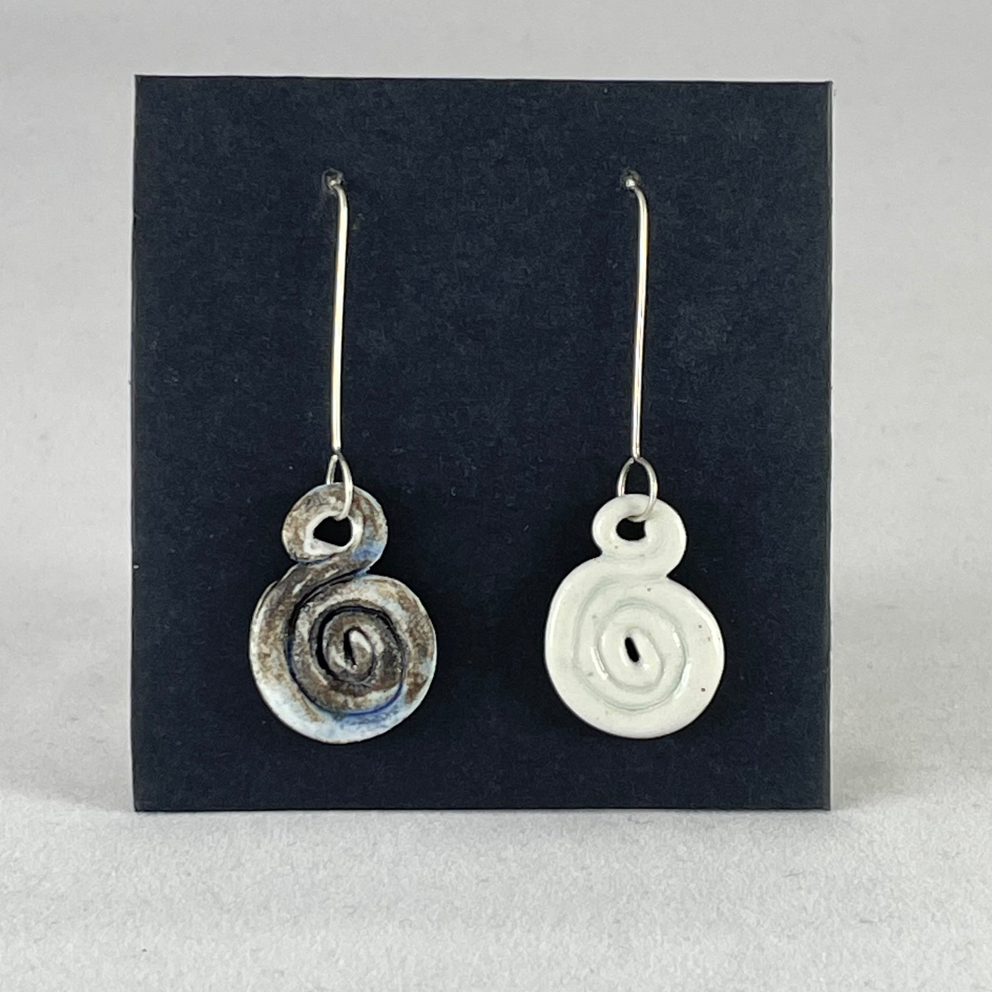 A pair of hand coiled porcelain earrings. One earring is white and the other is blue and black. Photographed on a black background. On sterling silver earrings hooks. 