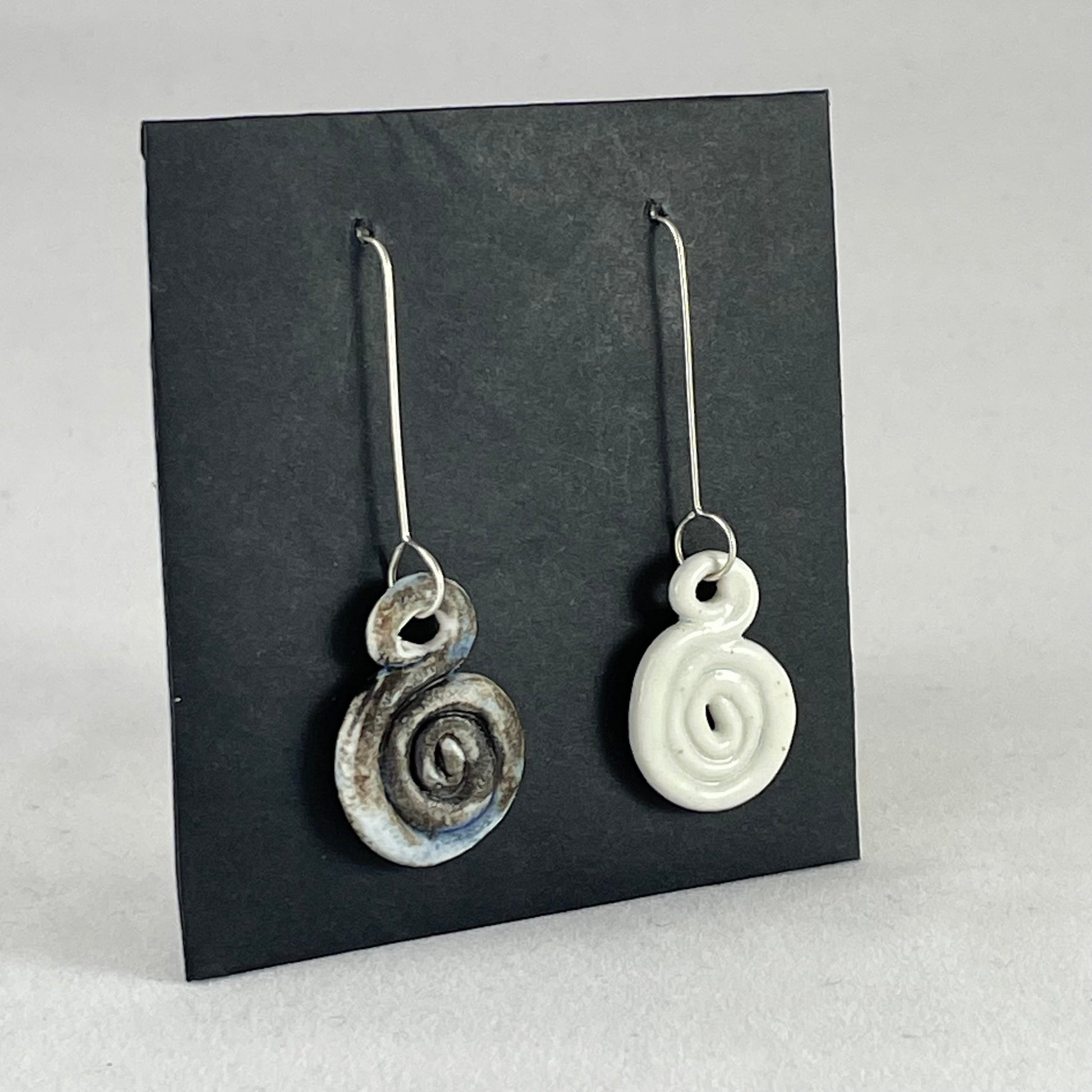 A pair of hand coiled porcelain earrings. One earring is white and the other is blue and black. Photographed on a black background. On sterling silver earrings hooks. Side view