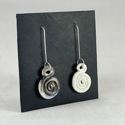 A pair of hand coiled porcelain earrings. One earring is white and the other is blue and black. Photographed on a black background. On sterling silver earrings hooks. Side view