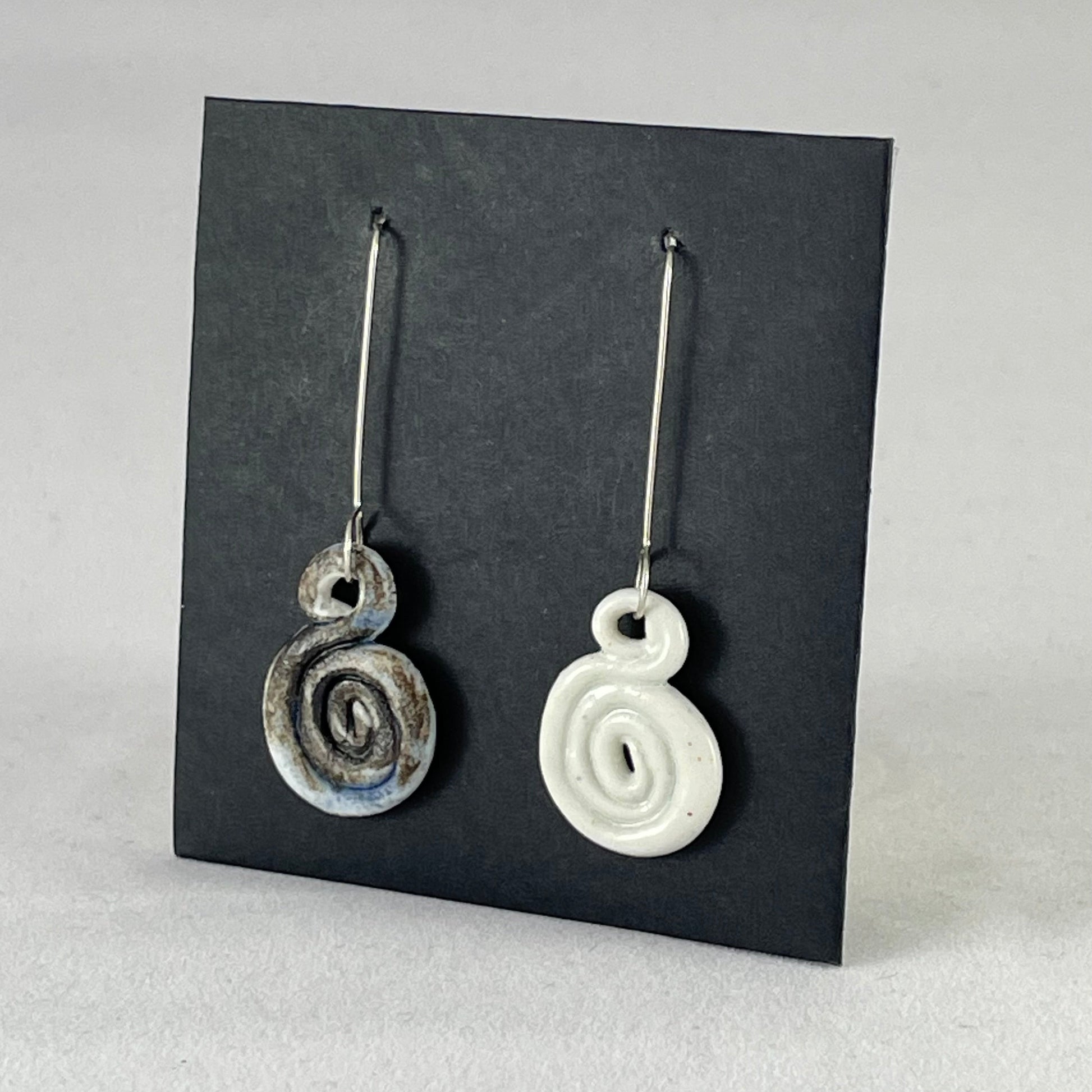 A pair of hand coiled porcelain earrings. One earring is white and the other is blue and black. Photographed on a black background. On sterling silver earrings hooks. Side view