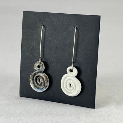 A pair of hand coiled porcelain earrings. One earring is white and the other is blue and black. Photographed on a black background. On sterling silver earrings hooks. Side view
