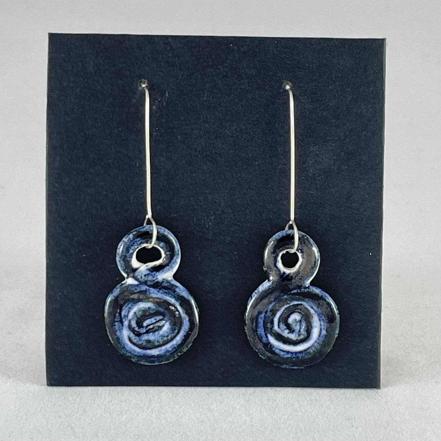 A pair of hand coiled porcelain earrings in dark blue. Photographed on a black background. On sterling silver earrings hooks.