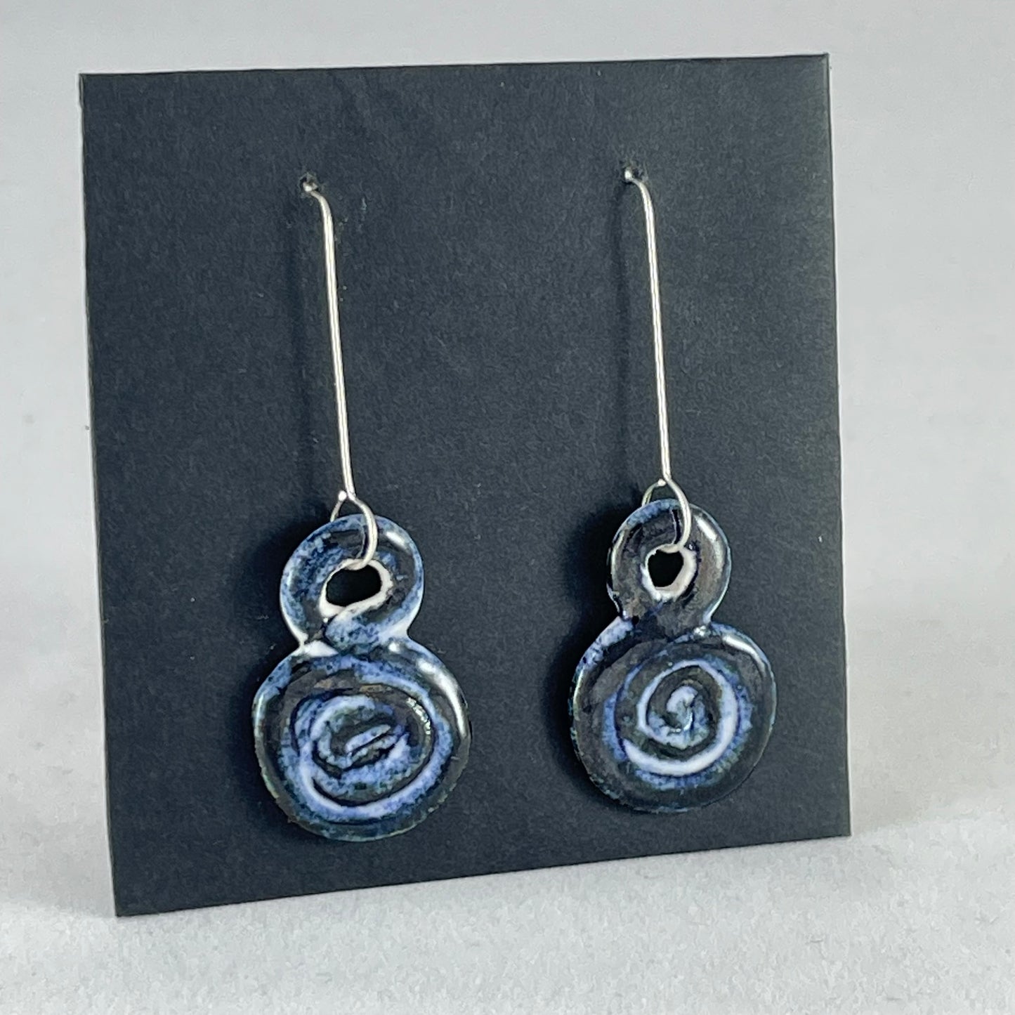 A pair of hand coiled porcelain earrings in dark blue. Photographed on a black background. On sterling silver earrings hooks. Side view