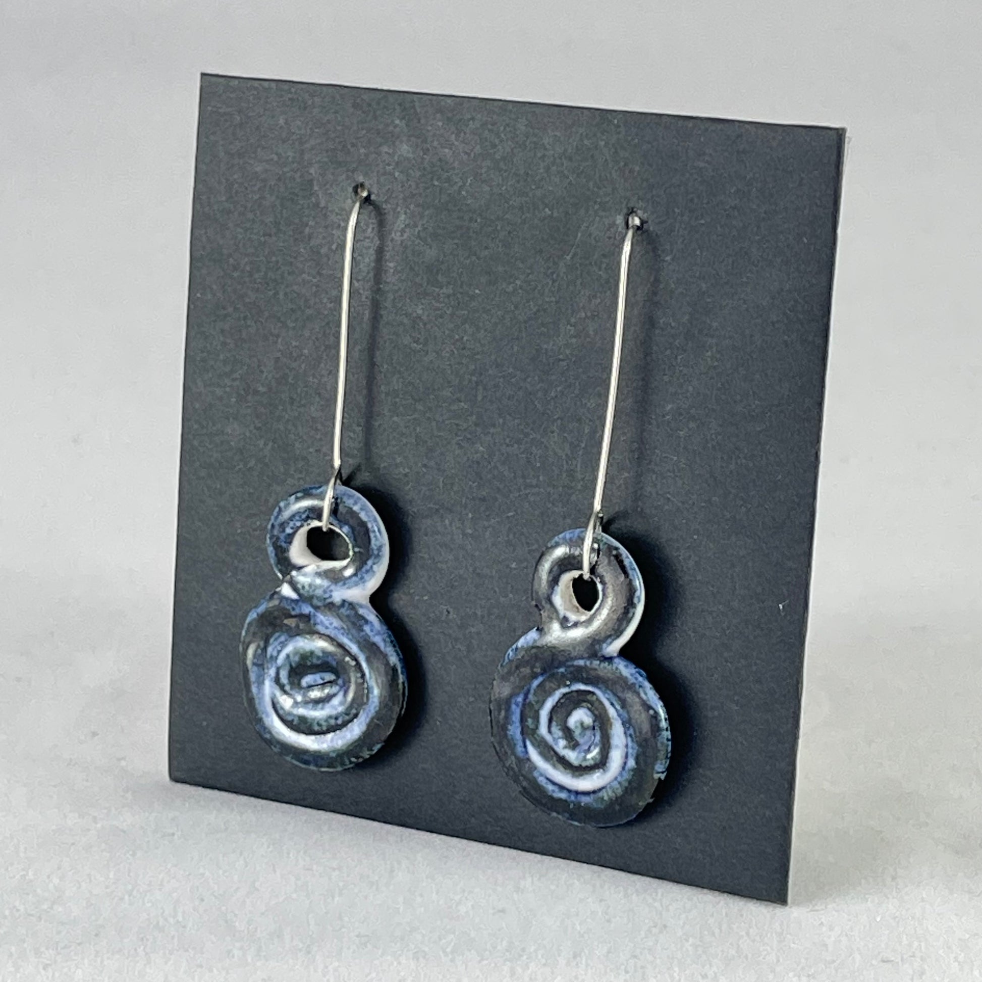 A pair of hand coiled porcelain earrings in dark blue. Photographed on a black background. On sterling silver earrings hooks. Side view