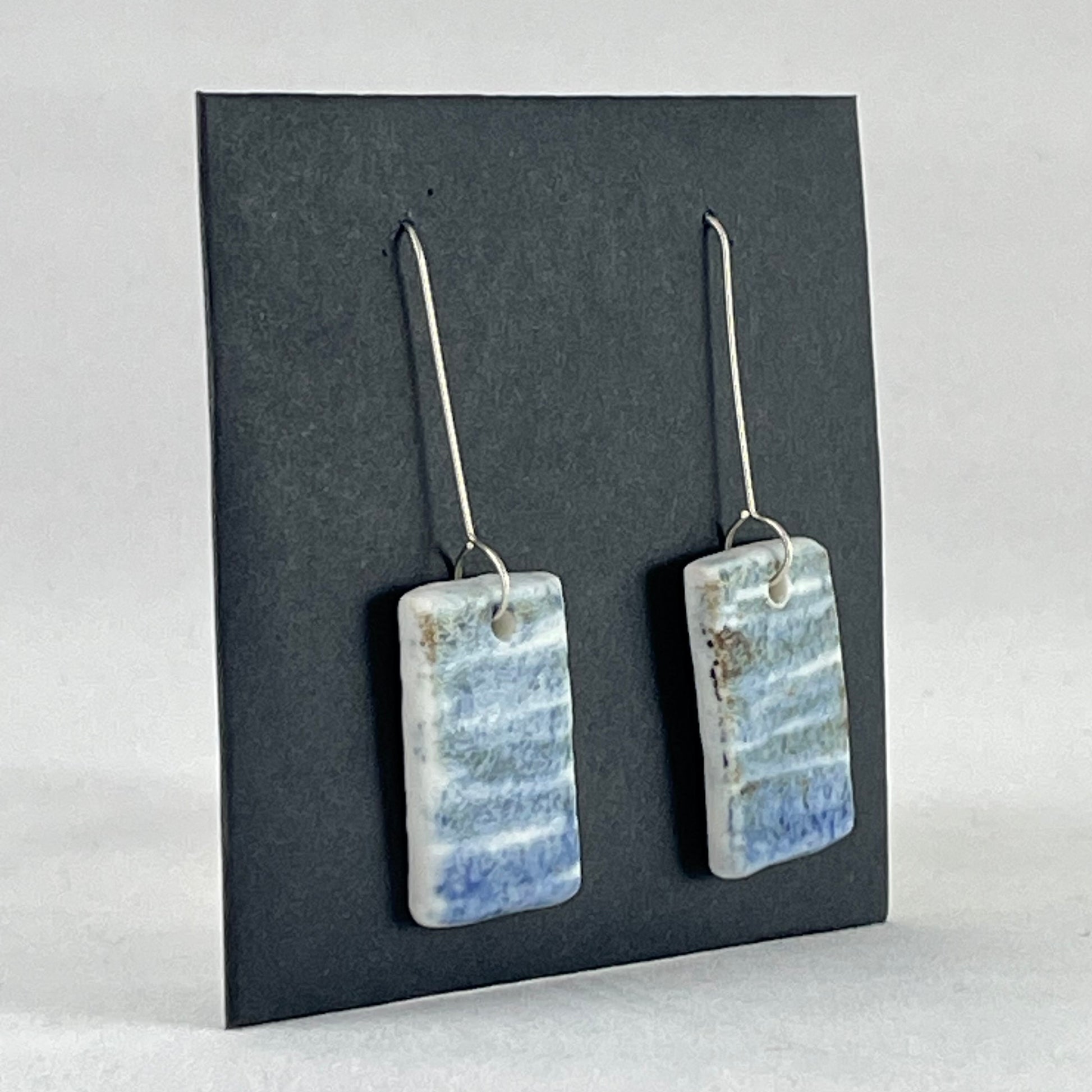 A pair of blue green and white porcelain earrings. Photographed on a black background. On sterling silver earrings hooks. Side view