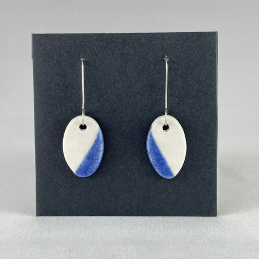 A pair of oval porcelain earrings in white with a diagonal strip of ultramarine blue. Photographed on a black background.