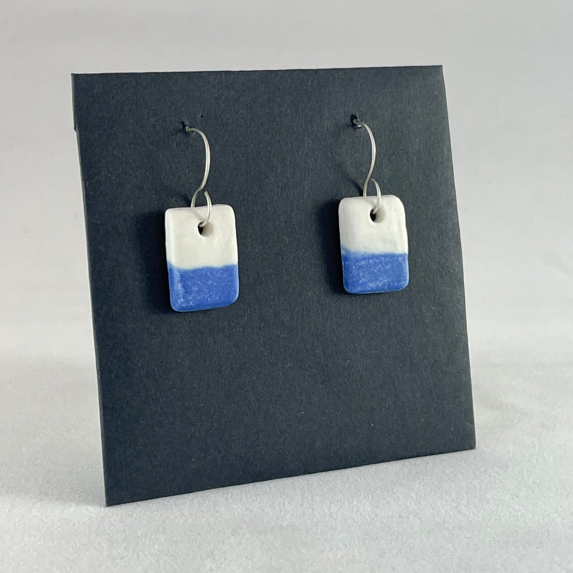 A pair of rectangular porcelain earrings in half white and half ultramarine blue. Photographed on a black background. Side view 