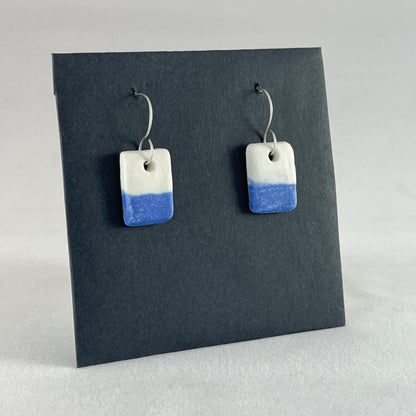 A pair of rectangular porcelain earrings in half white and half ultramarine blue. Photographed on a black background. Side view 