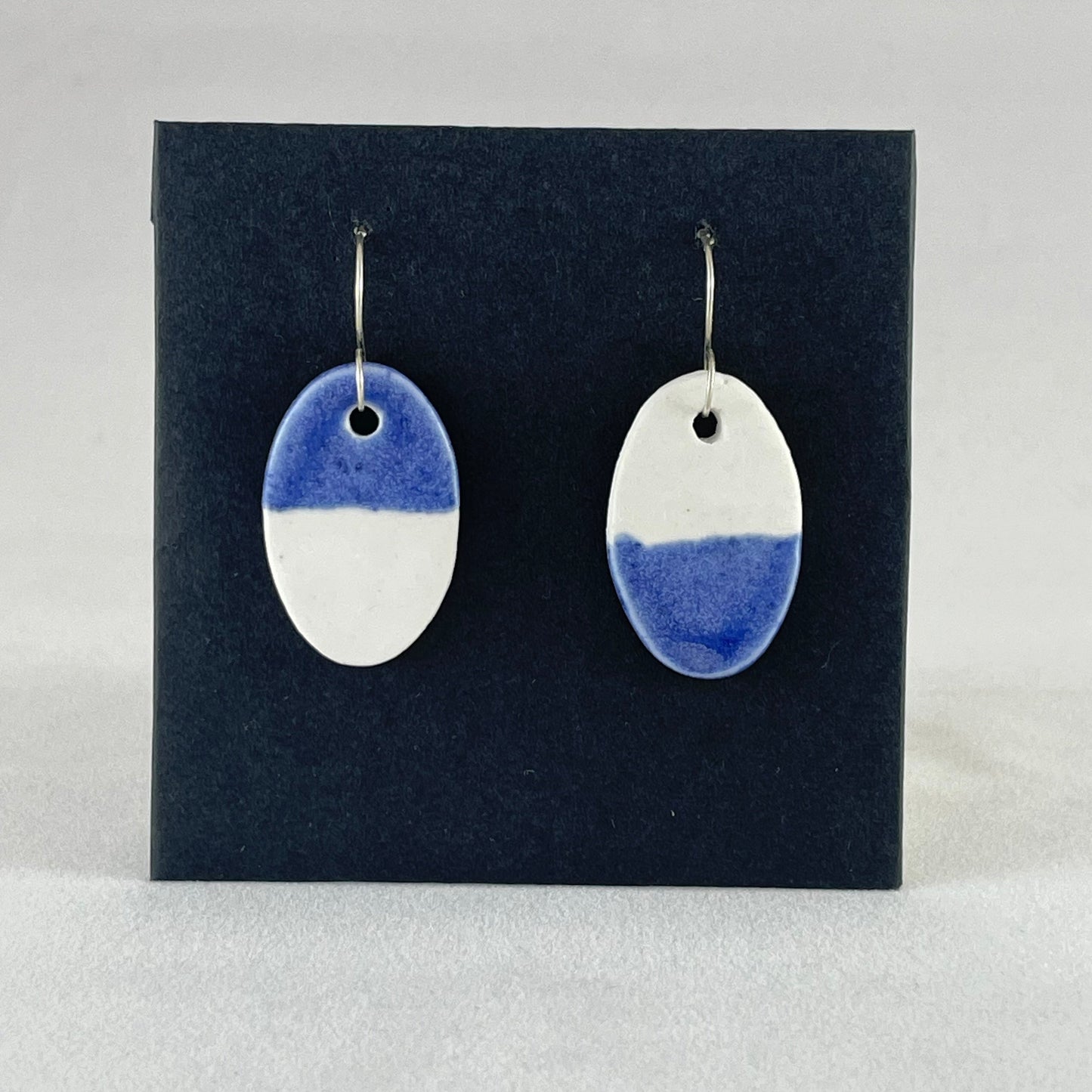A pair of oval shaped porcelain earrings in half white and half ultramarine blue. Photographed on a black background.