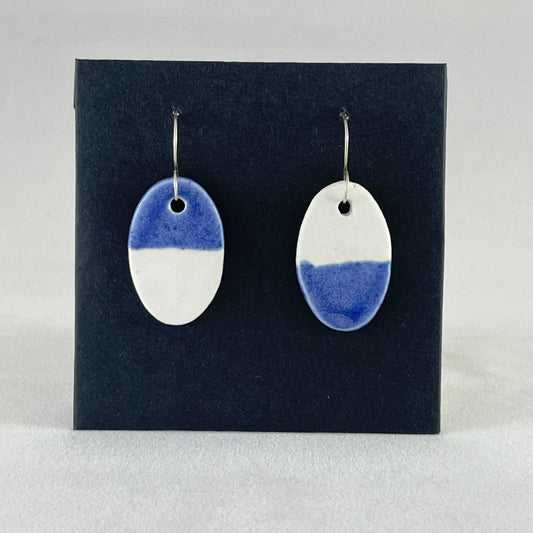 A pair of oval shaped porcelain earrings in half white and half ultramarine blue. Photographed on a black background.
