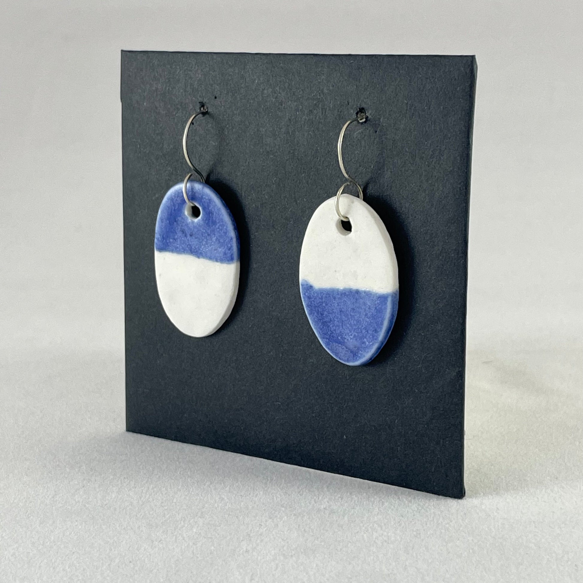 A pair of oval shaped porcelain earrings in half white and half ultramarine blue. Photographed on a black background. Side view