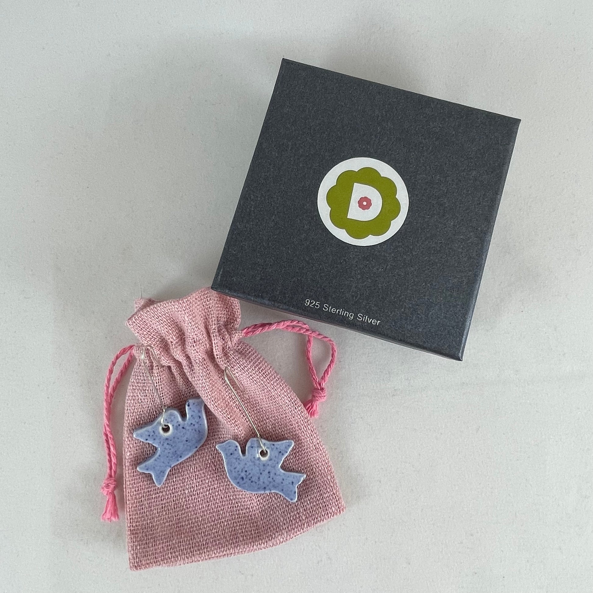 A pair of blue porcelain earrings in the shape of flying dove. Photographed on a linen pouch next to jewellery box.