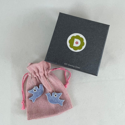 A pair of blue porcelain earrings in the shape of flying dove. Photographed on a linen pouch next to jewellery box.