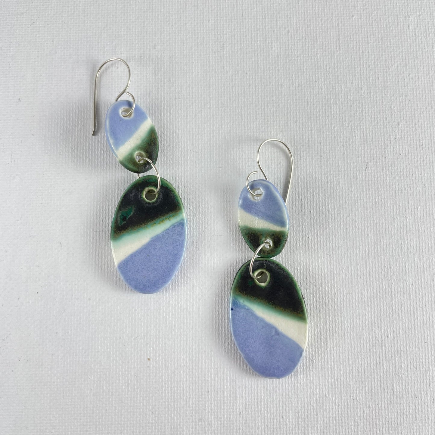 A pair of handmade porcelain earrings on a white background. Asymmetrical glaze pattern in very dark green, light blue and white on oval pendants.