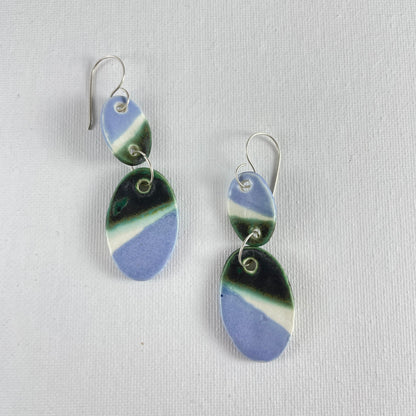 A pair of handmade porcelain earrings on a white background. Asymmetrical glaze pattern in very dark green, light blue and white on oval pendants.