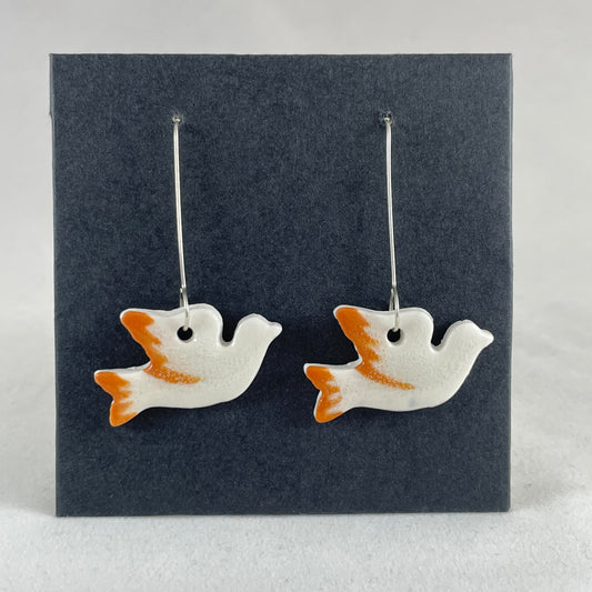 A pair of white and orange porcelain earrings in the shape of flying dove on black card.