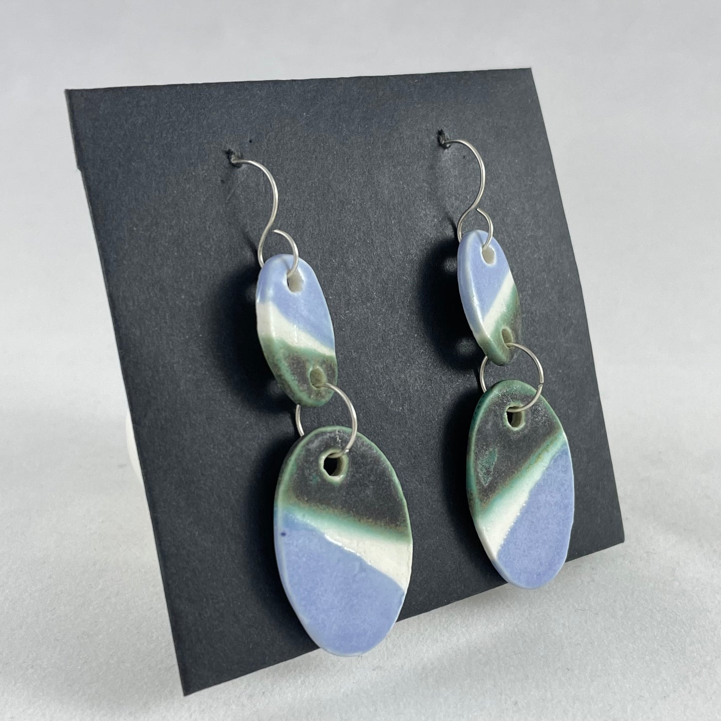 A pair of handmade porcelain earrings on a black background. Asymmetrical glaze pattern in very dark green, light blue and white on oval pendants. Side view (taken from left)