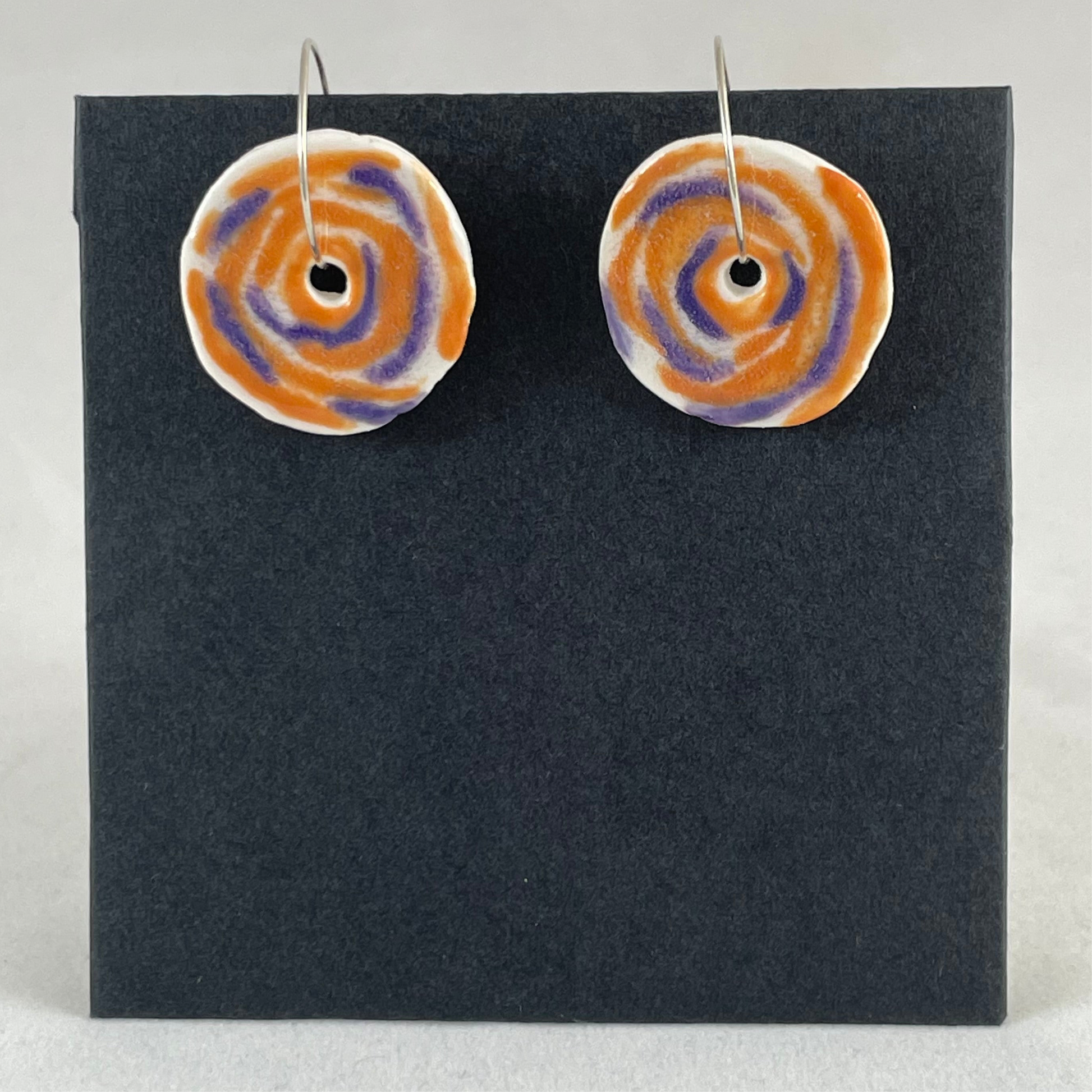A pair of lavender and orange porcelain earrings on black card. Simple rose pattern with loosely painted orange and lavender circles and small areas of white porcelain.