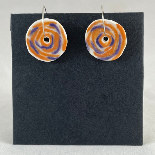 A pair of lavender and orange porcelain earrings on black card. Simple rose pattern with loosely painted orange and lavender circles and small areas of white porcelain.