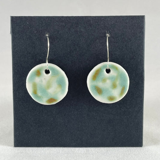 A pair of mint, olive green and white porcelain earrings hanging on black card. Splodges of glossy glaze look vaguely like a blurry camouflage pattern.