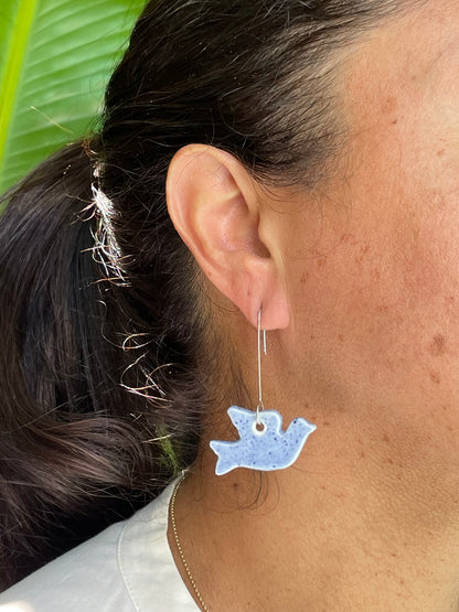 Closeup of woman wearing pale blue dove earrings. Woman has olive skin and her dark brown hair is pulled back in ponytail. Sun is shining in garden
