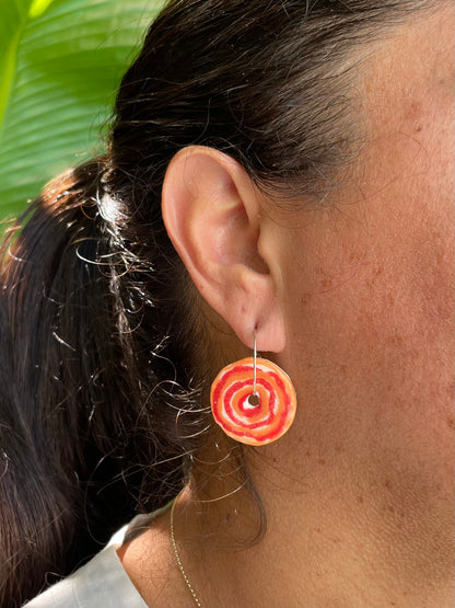 Closeup of woman wearing red and orange hoops earrings. Earrings are in a simple rose pattern with alternating orange and red circles painted loosely. Woman has tan skin and dark brown hair pulled back in a ponytail. A vivid green bird of paradise leaf is in the background and the sun is shining on her hair.