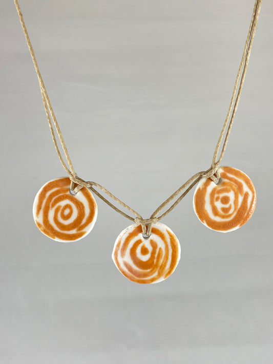 Necklace with three circular porcelain pendants on natural coloured thread, hanging in front of a white background. Orange rose pattern painted on white porcelain.  