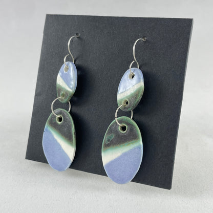 A pair of handmade porcelain earrings on a black background. Asymmetrical glaze pattern in very dark green, light blue and white on oval pendants. Side view (taken from right)