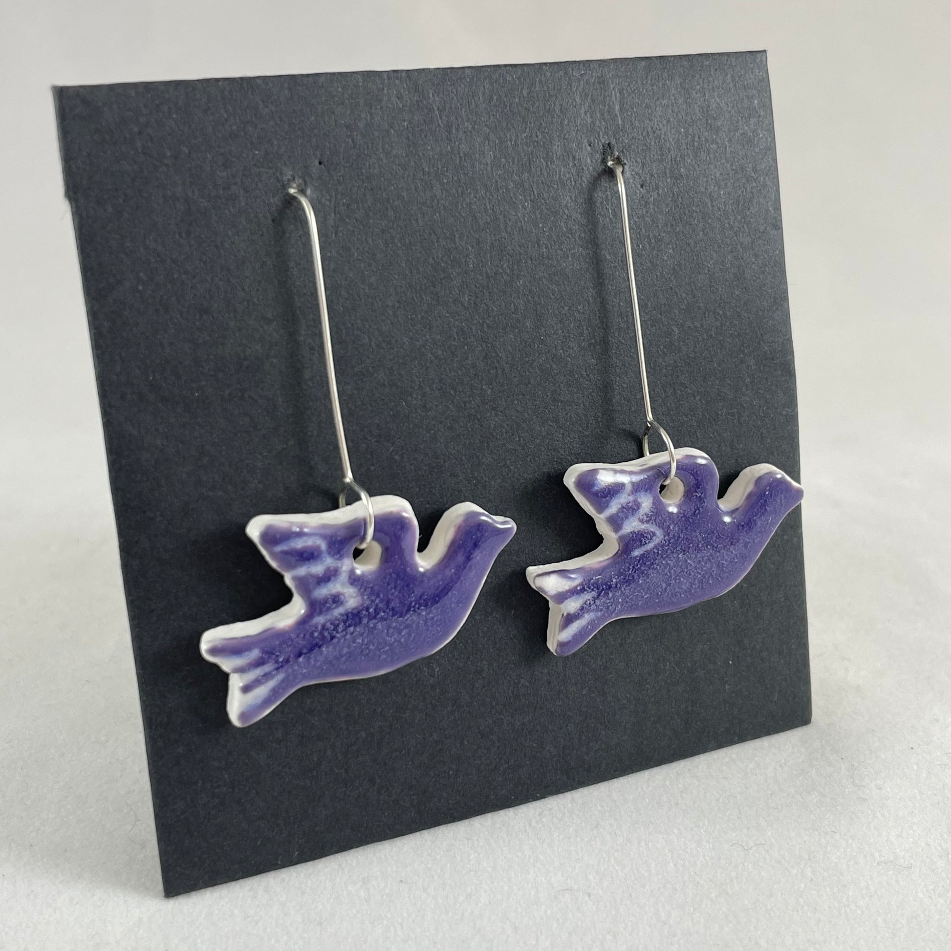 A pair of lavender porcelain earrings in the shape of flying dove on a black background. Side view