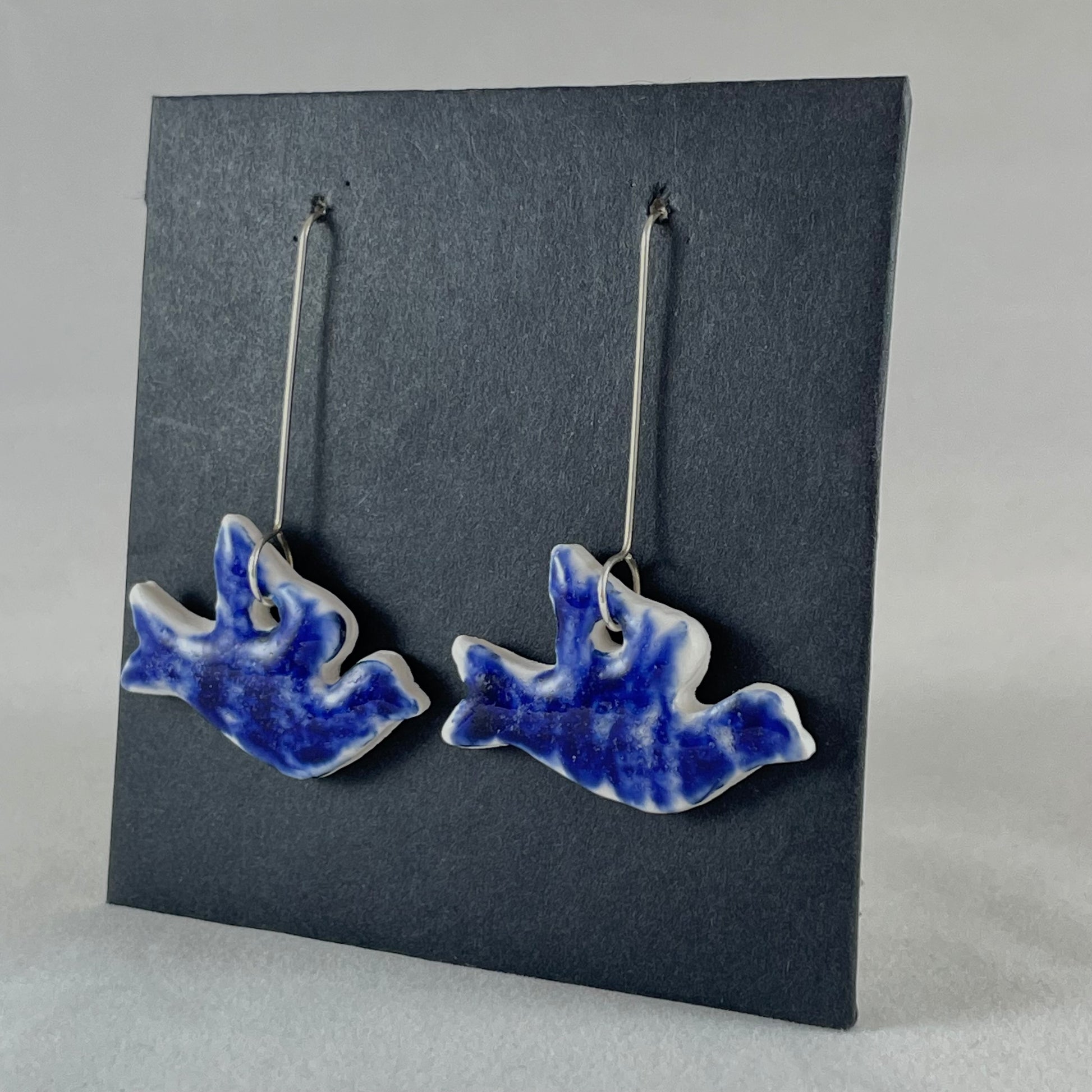 A pair of ultramarine blue porcelain earrings in the shape of flying dove on black card. Photographed on an angle from the right 