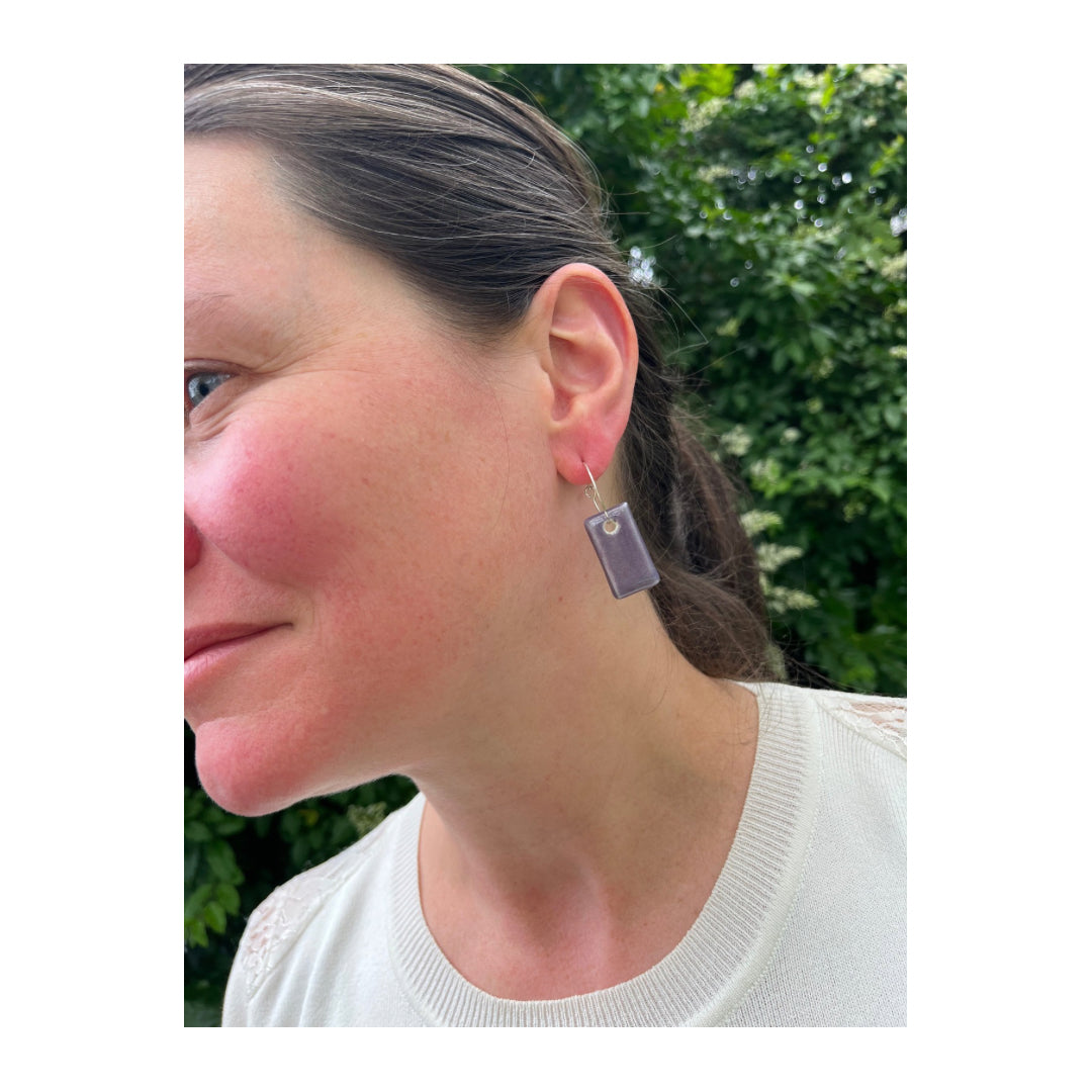 White woman with brown hair wearing purple pendant earrings on silver hooks. Looking into distance. One earring visible