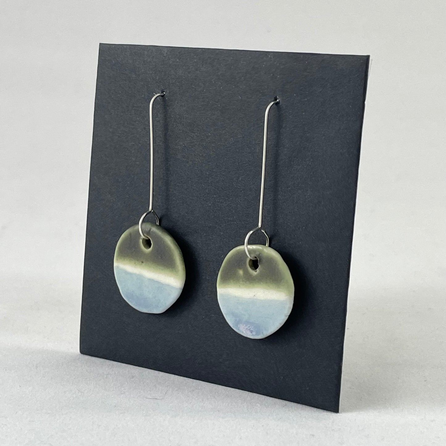Pair of circular porcelain earrings with  bands of colour: dark green, white and pale blue. On long sterling silver earring hooks. Side view