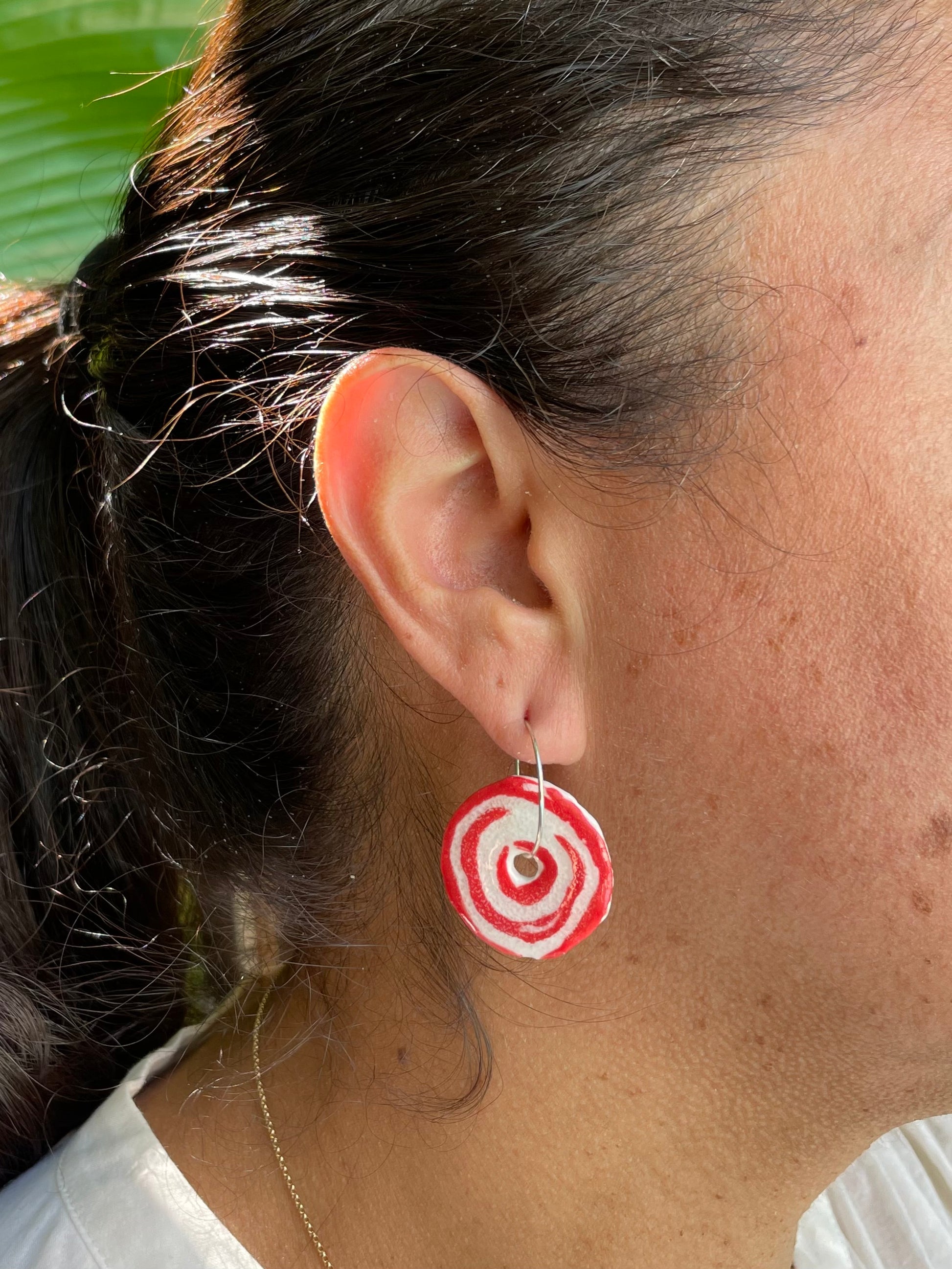 Closeup of woman wearing red and white hoops earrings. Woman has olive skin and her dark brown hair is pulled back in ponytail. Sun is shining on her hair