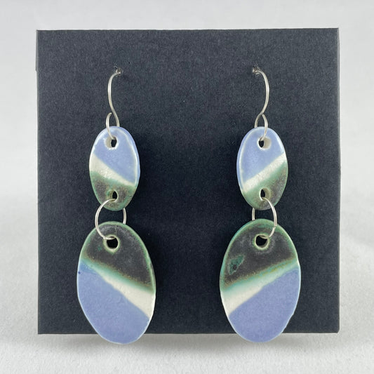 A pair of handmade porcelain earrings on a black background. Asymmetrical glaze pattern in very dark green, light blue and white on oval pendants.