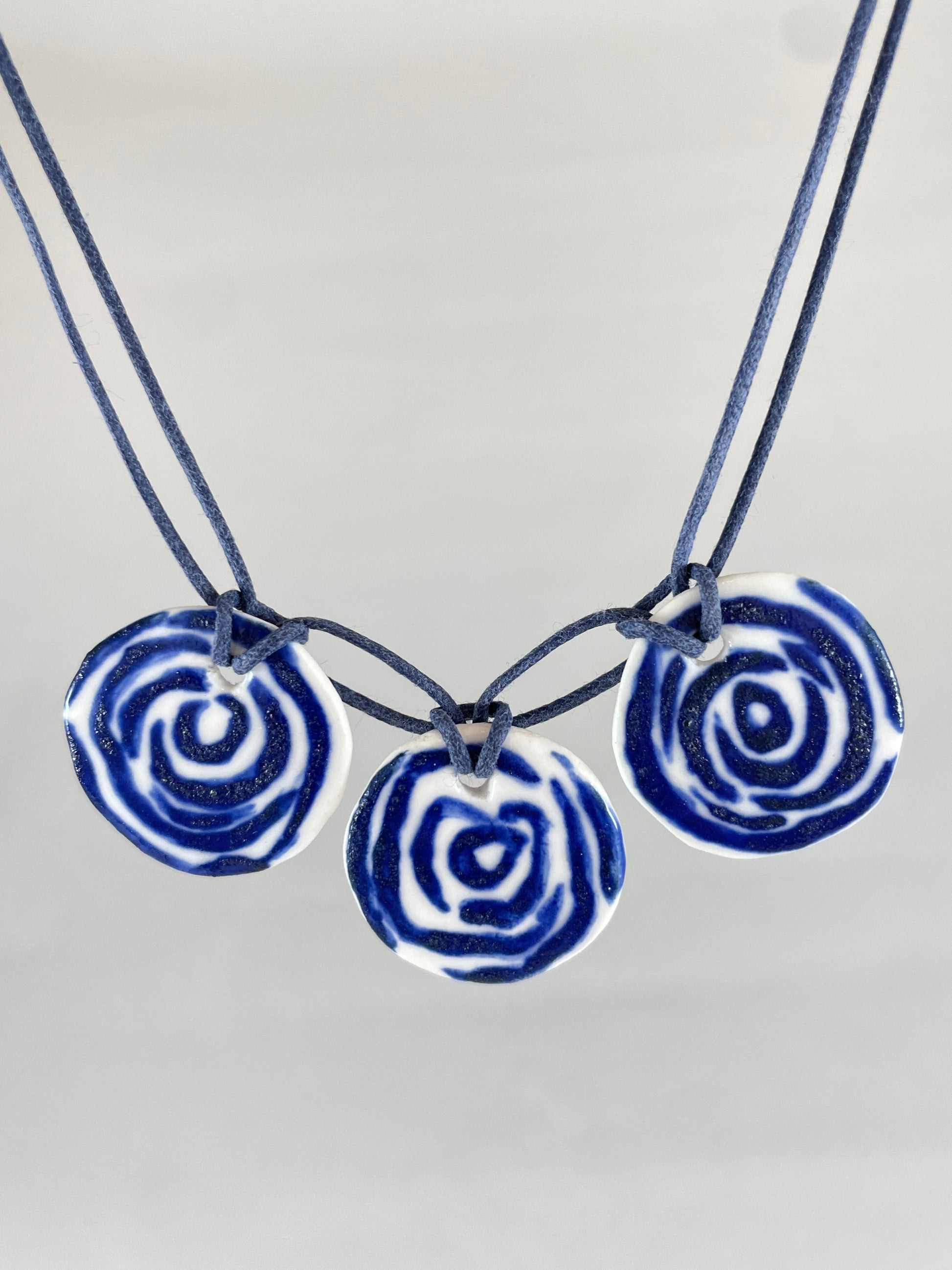 Closeup of necklace with three circular porcelain pendants on navy blue coloured thread, hanging in front of a white background. Blue rose pattern painted on white porcelain.  