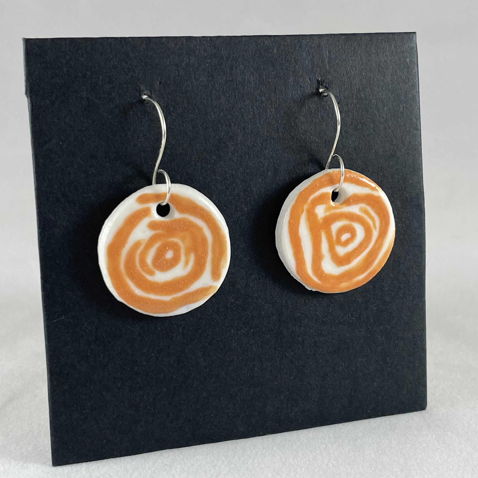 A pair of orange and white porcelain earrings hanging on black card. Loosely painted orange circles on white porcelain. Photographed from right hand side showing slim earring profile.