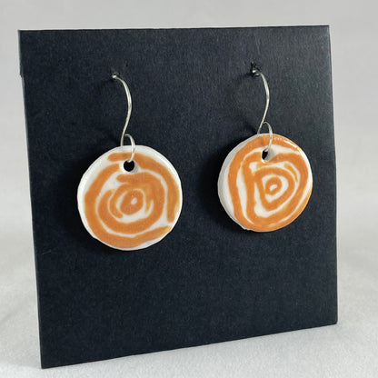 A pair of orange and white porcelain earrings hanging on black card. Loosely painted orange circles on white porcelain. Photographed from right hand side showing slim earring profile.