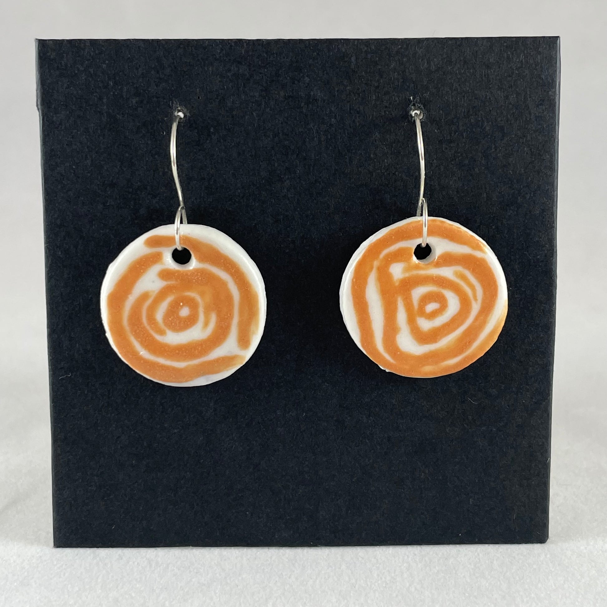 A pair of orange and white porcelain earrings hanging on black card. Simple rose pattern with loosely painted orange circles on white porcelain.