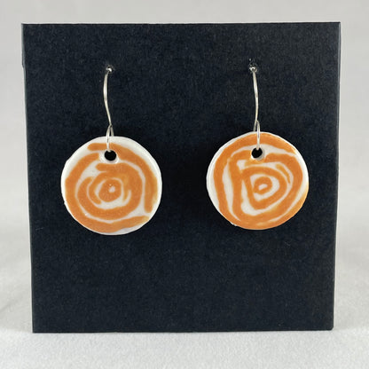 A pair of orange and white porcelain earrings hanging on black card. Simple rose pattern with loosely painted orange circles on white porcelain.
