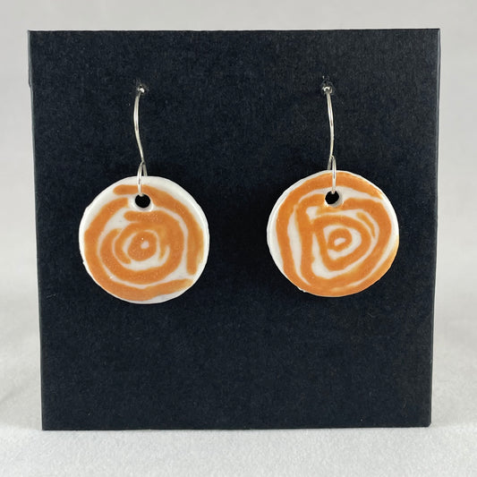 A pair of orange and white porcelain earrings hanging on black card. Simple rose pattern with loosely painted orange circles on white porcelain.