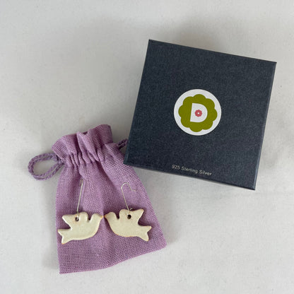 A pair of cream porcelain earrings in the shape of flying dove. Photographed on a linen pouch next to jewellery box.