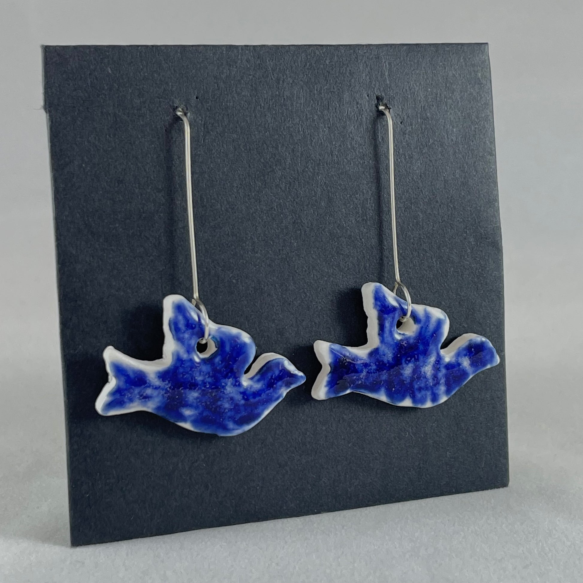A pair of ultramarine blue porcelain earrings in the shape of flying dove on black card. Photographed on an angle from the left 