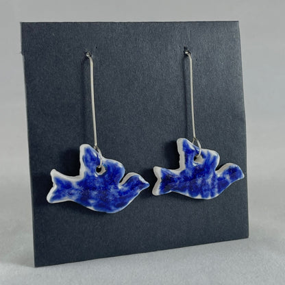 A pair of ultramarine blue porcelain earrings in the shape of flying dove on black card. Photographed on an angle from the left 