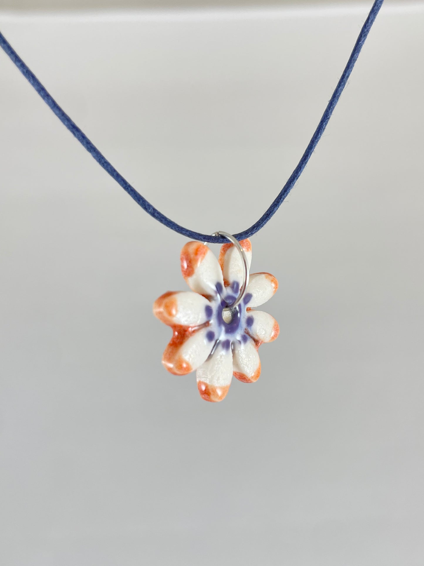 Necklace with one small daisy shaped porcelain pendant hanging on navy blue thread. Painted with lavender glaze in the centre and orange at the ends of petals. Photographed from left hand side showing thin pendant profile 